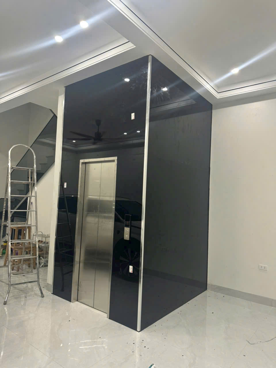 Thang máy gia đình cửa inox đang lắp đặt trong phòng khách hiện đại, cạnh thang nhôm và trần đèn LED